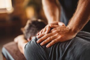 A person sits on an examination table while a chiropractor gently checks their neck and upper back.